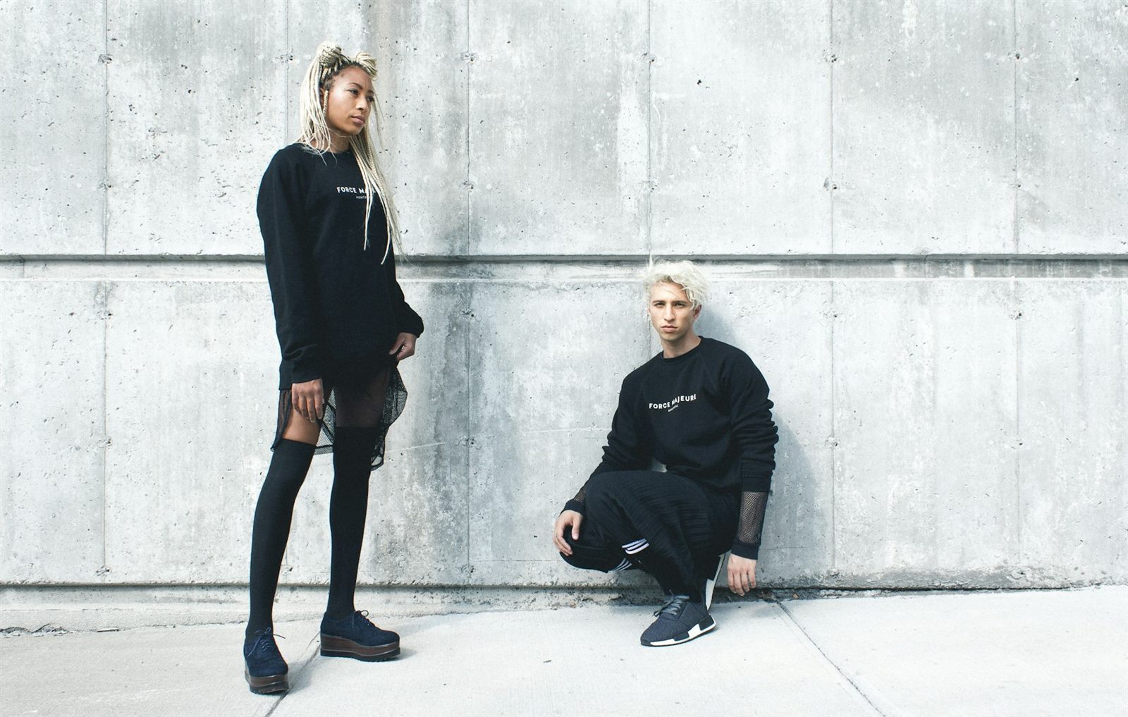 Two models in black crewneck sweatshirts against a grey concrete wall