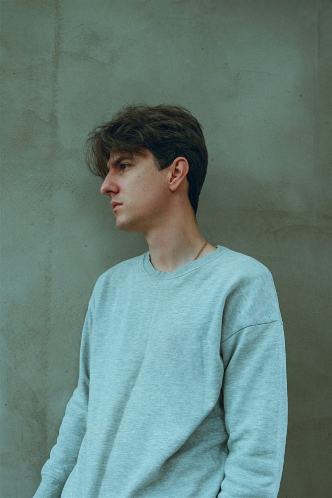 Young man in a pale blue crewneck sweatshirt against a grey-green wall