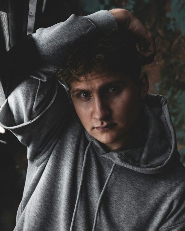Man in a grey hoodie leaning against a dark wall